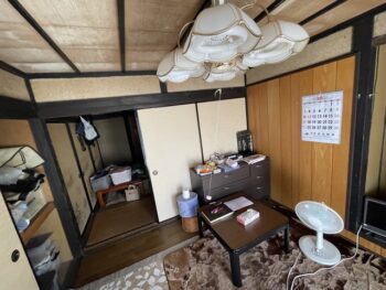 リフォーム前の和室別アングル。障子・エアコン・押入れの布団や荷物が積まれ、暗く閉塞感のある空間