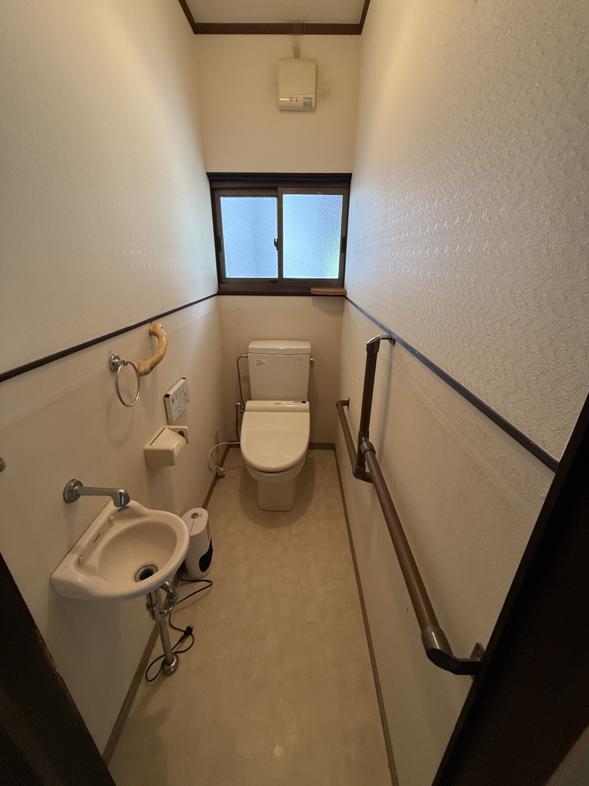 リフォーム前のトイレ。古いピンク系便器・凹凸壁紙・茶色T字手すり（安芸高田市K様邸）